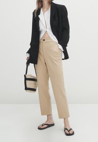 Beiget räätälöidyt housut, musta kaksirivinen bleiseri, valkoinen paita, mustat varvassandaalit, kädessä pieni mustilla yksityiskohdilla varustettu punottu laukku.