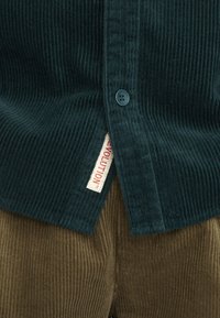 Donkergroene corduroy shirt met geribbelde textuur, voorzien van twee knopen en een label met "EVOLUTION." Gecombineerd met bruine corduroy broek.
