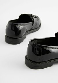 Pantofi din piele patentată neagră, cu formă rotundă, cusătură texturată și talpă joasă din cauciuc. Caracteristici un curea decorativă și cataramă.
