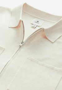 Vaalean beige vetoketjullinen takki, joka on valmistettu pehmeästä kankaasta. Takissa on kaulus ja kaksi etutaskua. Vetoketjussa on hopeinen metallinen vetotanko.