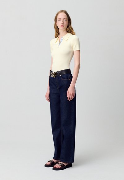 Camisa polo de rayas ribeteadas en amarillo pálido, ajustada, con cuello en punta, combinada con vaqueros de pierna ancha en denim oscuro y sandalias negras con detalles en dorado.