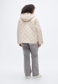 Prešita bež jakna s kapuco in ohlapnim krojem, z diamantnim vzorcem, elastiko na manšetah in ravnim robom. Kombinirana z sivimi hlačami in belimi supergami.