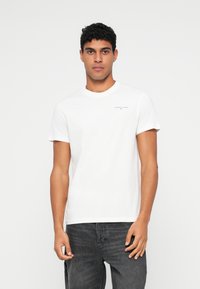 Νεαρός άνδρας με κοντά σγουρά μαλλιά, φορώντας ένα λευκό μπλουζάκι Tommy Jeans και σκούρο γκρι τζιν, που στέκεται μπροστά σε λευκό φόντο.