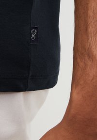 Marineblaue T-Shirt mit weicher Textur und kurzen Ärmeln, ausgestattet mit einem kleinen Markierungslabel nahe dem Saum. Weiße Stoff sichtbar darunter.