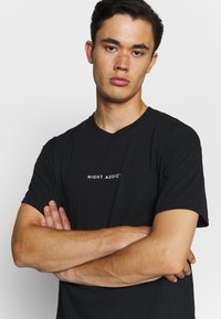 Juodas medvilninis marškinėliai su apvaliu iškirpimu ir trumpomis rankovėmis, su baltu užrašu "NIGHT ADDICT" krūtinėje.