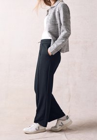 Giacca grigia texturizzata, top bianco, pantaloni larghi neri e sneakers bianche con accenti grigi. Tasche visibili nei pantaloni.