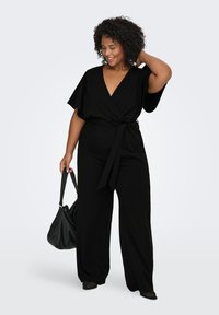 Juodas kombinezonas su plačiomis rankovėmis ir surišama juostele. Lygus audinys, V formos iškirptė ir plačių kojų stilius. Papuoštas juoda rankine.