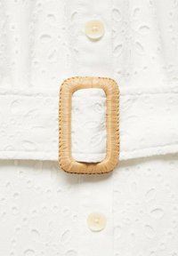 Gros plan sur un tissu blanc avec des motifs floraux brodés, des boutons crème et une boucle de ceinture tissée rectangulaire.