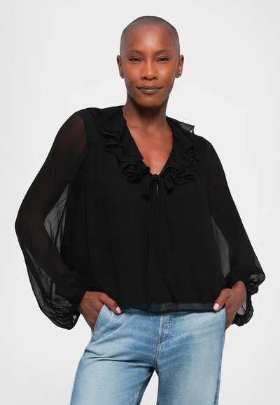 Zwarte blouse met franje bij de halslijn en mouwen, gemaakt van doorschijnende stof. Gekleed met lichtblauwe jeans, met een losse pasvorm en zakken.