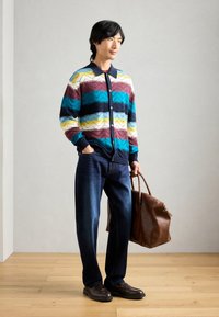 Cardigan à col pour homme avec des motifs zigzag multicolores en bleu, moutarde et bordeaux, associé à un jean en denim foncé et un sac en cuir marron.