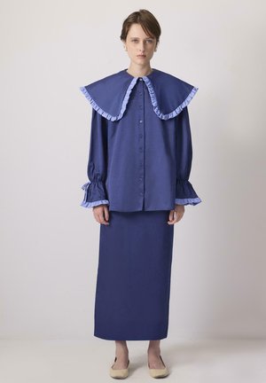 Una giovane donna indossa un abito blu con un ampio colletto a ruches e maniche lunghe, con le mani incrociate ai fianchi, su uno sfondo neutro.