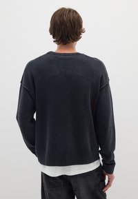 Maglione a maglia grigio scuro con orlo e polsini a coste. Presenta una vestibilità rilassata e cuciture visibili. Indossato sopra una camicia bianca a strati.