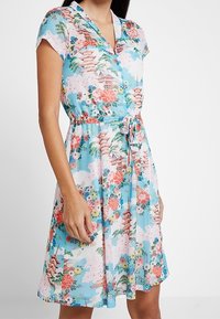 Blommönstrad klänning med ljusblå bakgrund, korta ärmar, V-ringning och bältesmidja. Innehåller mönster av pagoder och blommor.