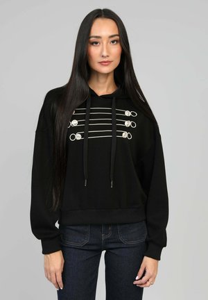 Hoodie noir avec des accents en métal argenté et des cordons épais, présentant des lignes horizontales et des boucles sur le devant. Porté avec un jean sombre.
