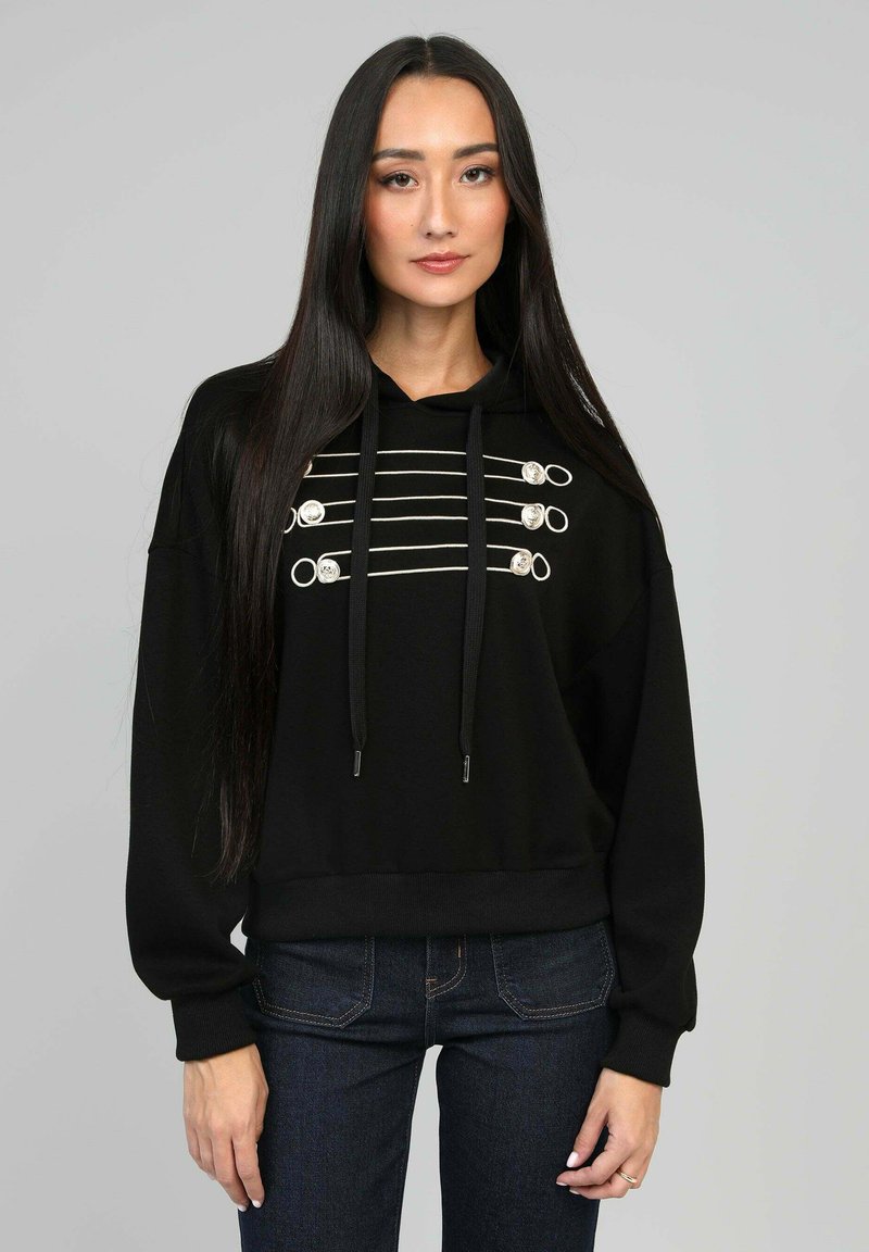Hoodie noir avec des accents en métal argenté et des cordons épais, présentant des lignes horizontales et des boucles sur le devant. Porté avec un jean sombre.