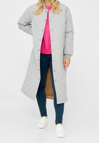 Persona che indossa un lungo cappotto trapuntato grigio chiaro aperto, che rivela una felpa rosa brillante, jeans blu scuro e sneakers bianche, in piedi contro uno sfondo bianco.
