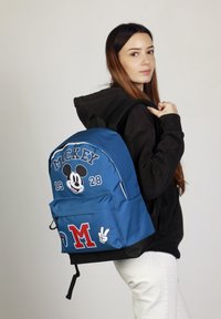 Mochila de tela azul con un gráfico de Mickey Mouse, texto "MICKEY" y el número "28". Incluye parches rojos con la "M" y el símbolo de paz.