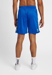 Blå träningstights med en texturerad tygkvalitet, prydda med ett gult sidomönster och ett elastiskt midjeband. I kombination med vita strumpor och sneakers.