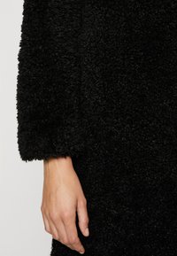 Cappotto nero e peloso con una texture morbida, caratterizzato da una manica cropped e rilassata e una finitura delle cuciture pulita. Mano visibile sul bordo.