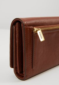 Brun læderpung med lynlås, der har fin stitching, en hardware tag i guldfarve og en struktureret, rektangulær form.