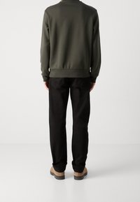 Dark green sweatshirt with ribbed cuffs and hem, paired with black pants and beige shoes, viewed from the back against a neutral background.