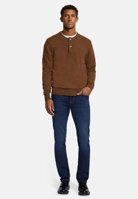 Maglione marrone a girocollo con pattina di bottoni, abbinato a jeans blu scuro e scarpe nere. Il maglione presenta un tessuto strutturato e polsini a costine.