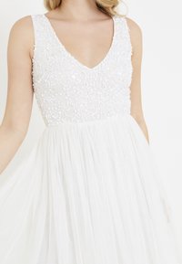 Valkoinen mekko, jossa on V-aukko, ja se on varustettu helmillä koristellulla yläosalla sekä virtaavalla, kerroksellisella tyllihameella. Pintakäsitellyt paljetit peittävät yläosan.