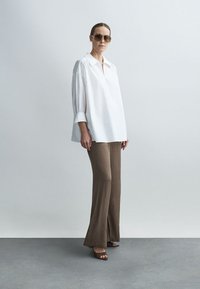 Blusa blanca de corte holgado con mangas largas; pantalones marrones texturizados y acampanados; combinados con gafas de sol marrones y sandalias de tacón ancho.