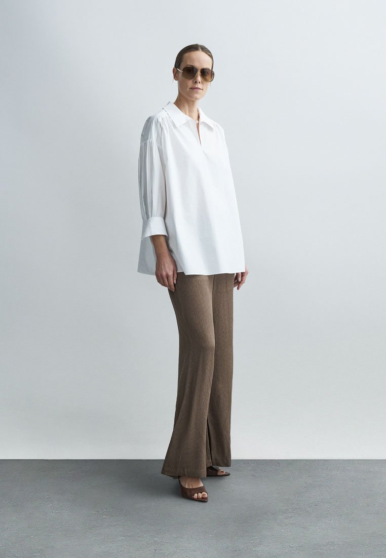 Blouse blanche ample à manches longues ; pantalon évasé marron texturé ; associé à des lunettes de soleil marron et des sandales à talons blocs.