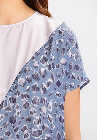 Blusa azul con una tela suave y ligera, con un cuello beige en contraste. Impresa con patrones abstractos oscuros, de manga corta y de corte holgado.