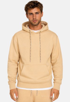 Homme aux cheveux courts bruns et aux yeux bleus portant un sweat à capuche beige et un pantalon assorti, se tenant devant un fond clair uni.
