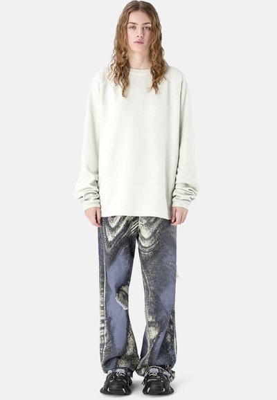 Joven con cabello rizado que lleva una sudadera extragrande color marfil, vaqueros holgados con lavado ácido y zapatillas negras, de pie contra un fondo blanco.