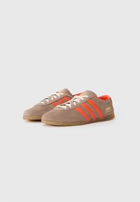 Tan mockasnike Adidas Gazelle med röda ränder, vita snören och gummifärgade sulor på vit bakgrund.