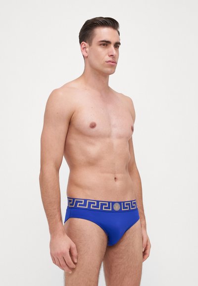 Blaue Schwimmbrieftaschen aus dehnbarem Stoff. Mit einem goldfarbenen griechischen Schlüsselmotiv am Bund und einem runden Logo-Akzent.