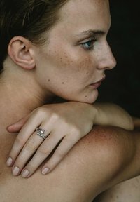 Silverring med en rosa sten, staplad design. Modellens hand vilar på axeln och visar upp slät hud med fräknar. Neutral bakgrund.