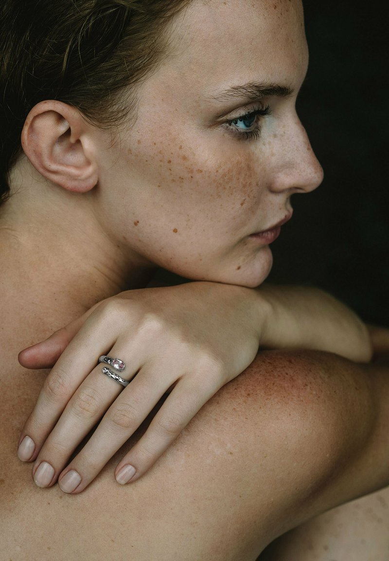 Silverring med en rosa sten, staplad design. Modellens hand vilar på axeln och visar upp slät hud med fräknar. Neutral bakgrund.