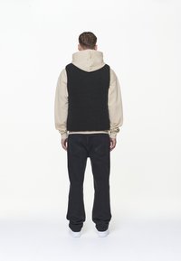 Svart, strukturerad väst över en beige hoodie, kombinerad med mörka jeans och vita sneakers. Vesten har en ärmlös design och en avslappnad passform.