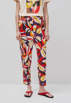 Donna con pantaloni colorati a motivo foglie, t-shirt grafica beige e sandali rossi in piedi su uno sfondo chiaro e semplice.