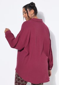Camicia oversize bordeaux realizzata in tessuto testurizzato, dotata di colletto, maniche lunghe e orlo arrotondato. Indossata con pantaloni fantasia.