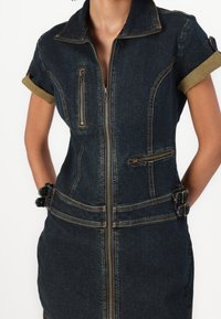 Mörk denimklänning med kort ärm och framdragkedja, flera dragkedjefickor, upprullade ärmslut och dubbla spännen i midjan.