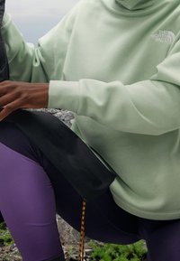 Ljusgrön sweatshirt med hög hals och logotyp. Svarta byxor med accentdetaljer, sittande på stenig mark med grön växtlighet.