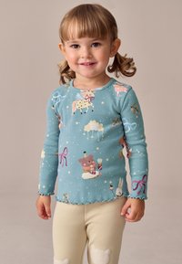 Világoskék bordázott hosszú ujjú ing, amelyen játékos állat- és felhőminták láthatók. Szegélye hullámos, puha tapintású, bézs leggingssel kombinálva.