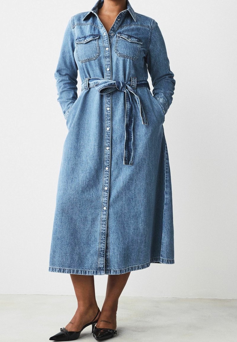 Femme portant une robe midi en denim à manches longues, boutonnée sur le devant, ceinturée, avec des poches poitrine et des escarpins noirs à bout pointu.