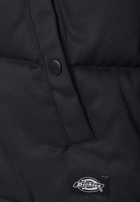 Doudoune noire avec un bouton brillant, un tissu texturé et une étiquette de marque visible avec un logo circulaire près de la couture.