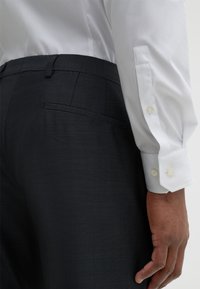 Schwarze, strukturierte Hose mit glattem weißem Hemd. Das Hemd hat geknöpfte Manschetten. Auf der Hose ist eine Gesäßtasche sichtbar.