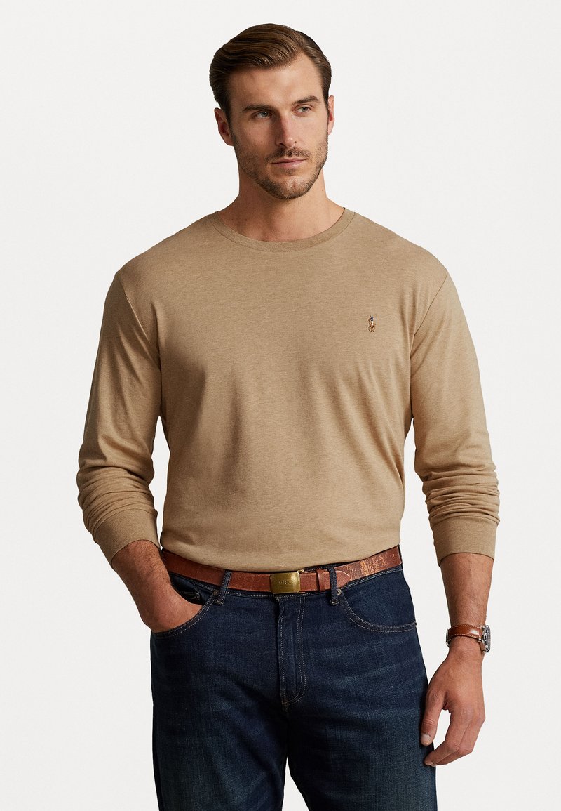 Beige μακρυμάνικη μπλούζα με στρογγυλή λαιμόκοψη, συνδυασμένη με σκούρα τζιν παντελόνια και καφέ ζώνη με μεταλλική αγκράφα. Διακριτική λεπτομέρεια λογότυπου.