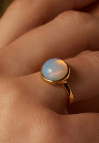 Goldring mit einem runden, hellblauen Stein von zarter Ausstrahlung, auf einem glatten, schmalen Band montiert. Strukturierte Details sind auf der Steinoberfläche sichtbar.