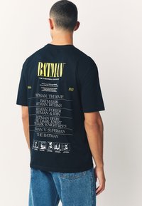 Camiseta negra de algodón con texto amarillo que dice "BATMAN" y una lista de películas desde 1966 hasta 2022 en la parte trasera, con detalles gráficos debajo.