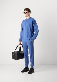 Blue long-sleeve sweatshirt and matching joggers in cotton blend, paired with black sneakers and a black duffel bag with shoulder straps.