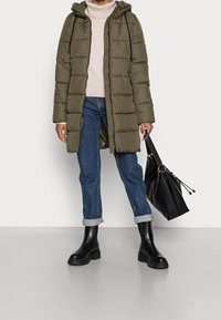 Giacca a vento verde oliva con cappuccio, chiusura con zip e tasche frontali. Abbinata a jeans chiari, stivali neri alla caviglia e una borsa nera con cordoncino.